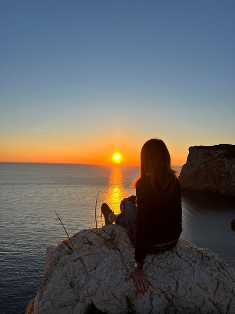 Donna seduta su una scogliera al tramonto che osserva l’orizzonte, simbolo di introspezione e viaggio interiore